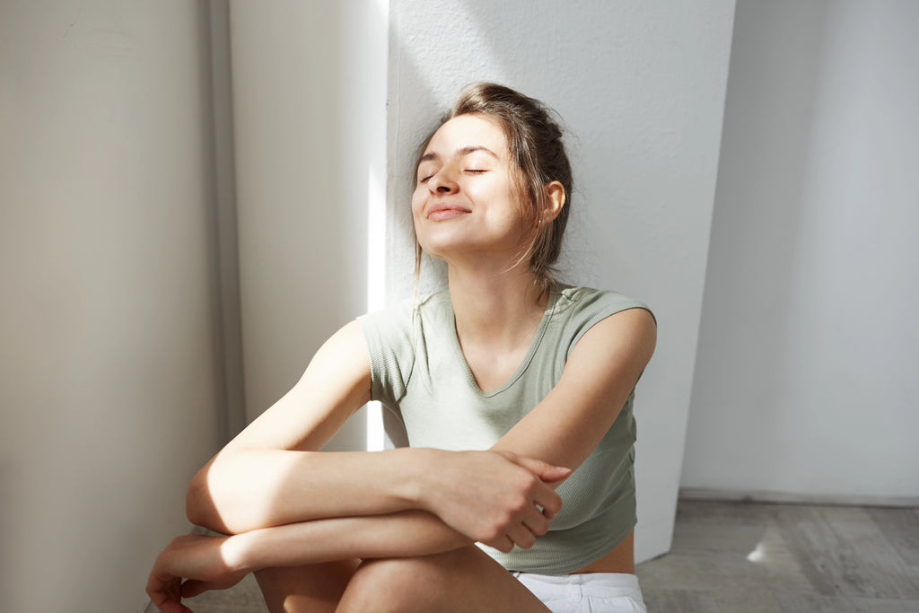 Portrait of a young woman smiling with closed eyes, enjoying morning sunlight while sitting on the floor by a white wall.